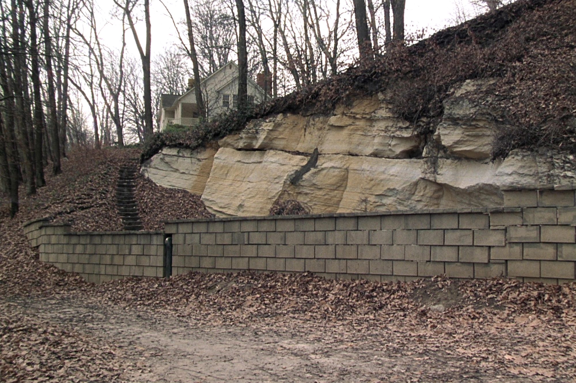 A block retaining wall at the foot of a limestone bluff, a house at the top of the wooded slope.