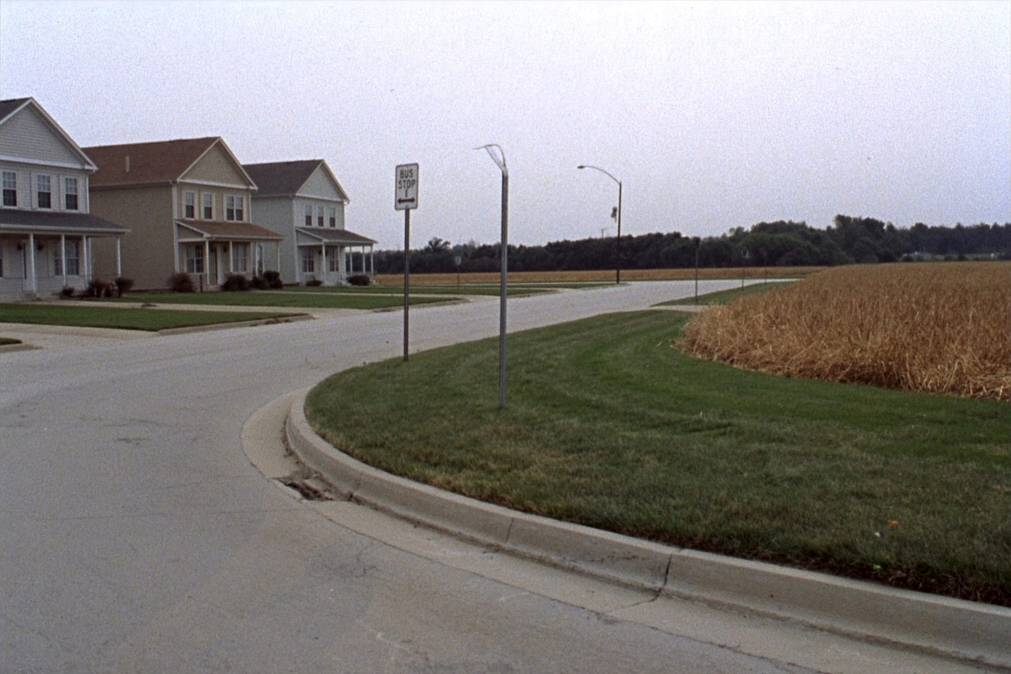 A suburban street ending mid-block where a harvested field begins, a bus stop sign at the curb.