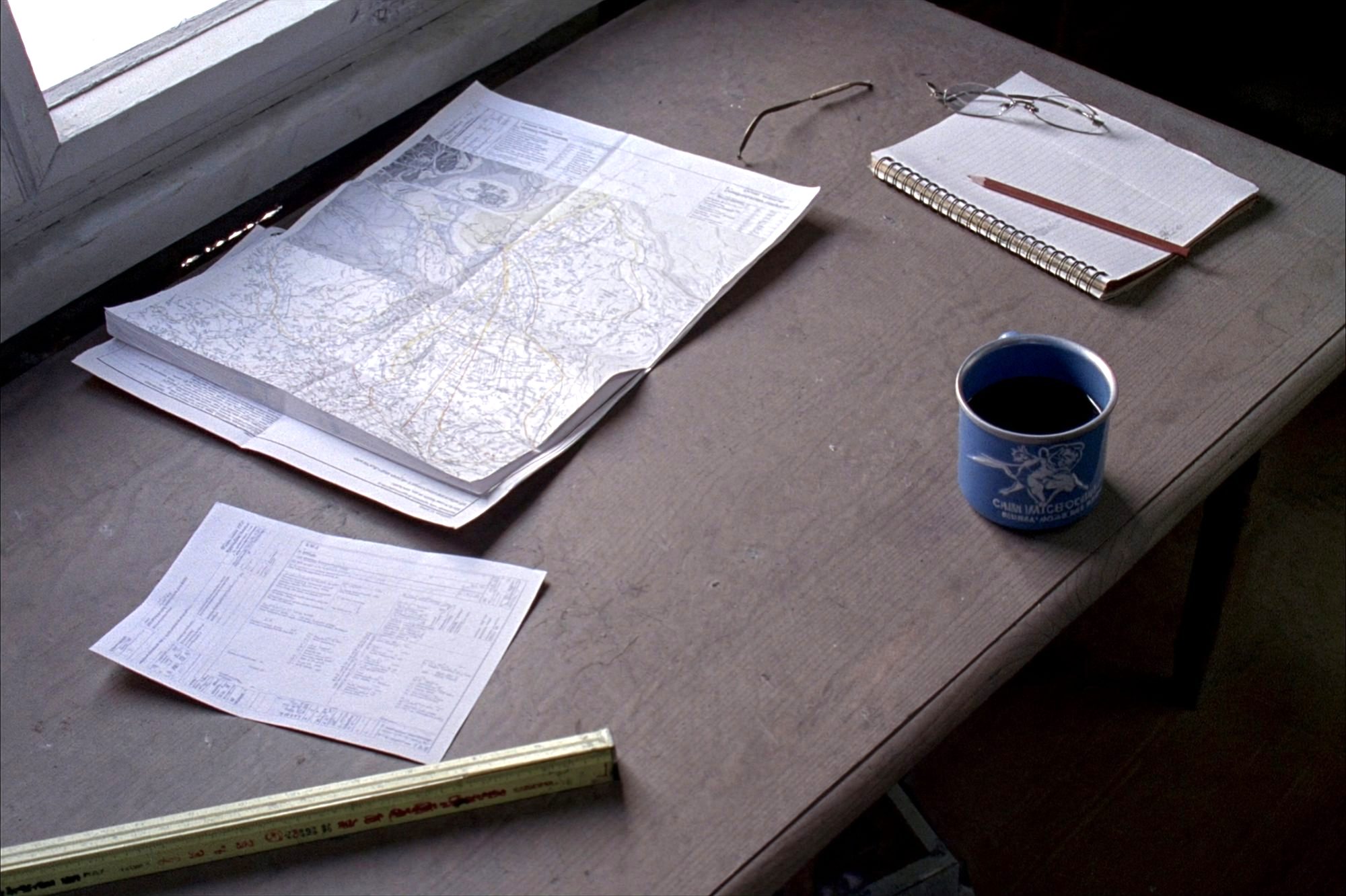A plain table in window light holding a folded plat map, a record sheet, a notebook and a coffee mug.